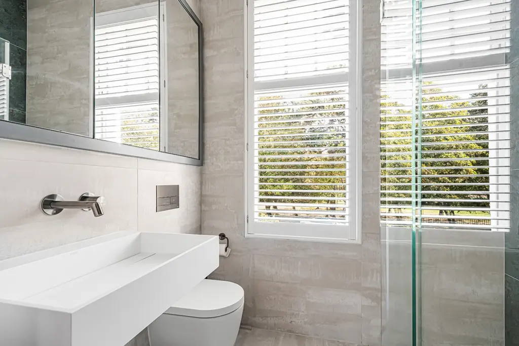 A modern bathroom featuring a white wall-mounted basin, a toilet, a mirrored cupboard, and a glass-enclosed shower, with windows covered by white shutters.