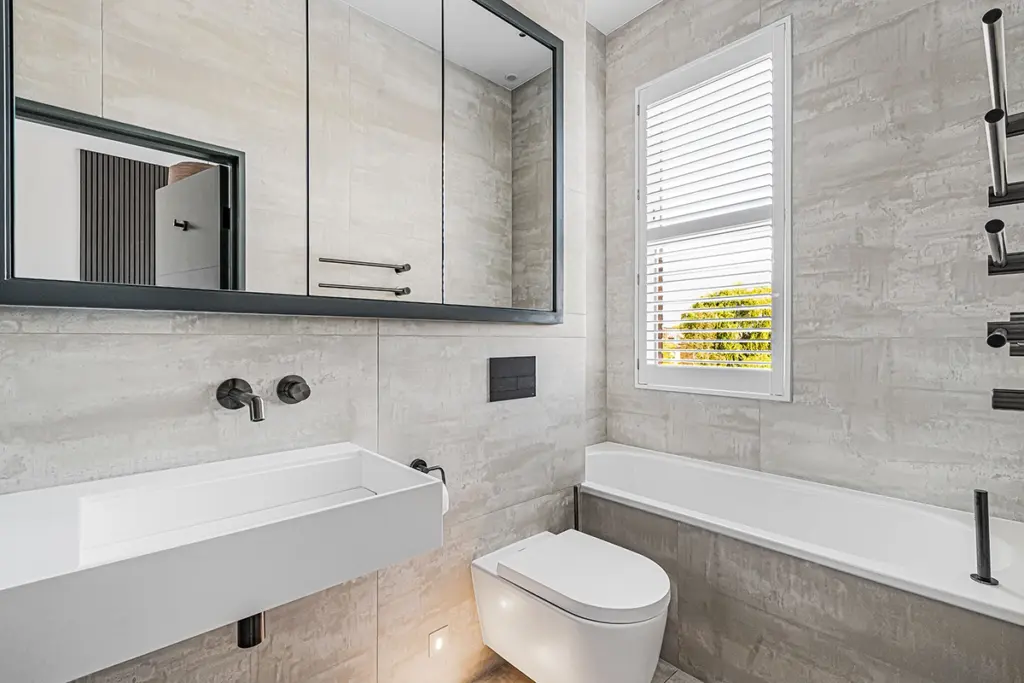 A modern bathroom with light-toned tiled walls, a wall-mounted white basin, a wall-hung toilet, a built-in bath, and a window with white shutters.