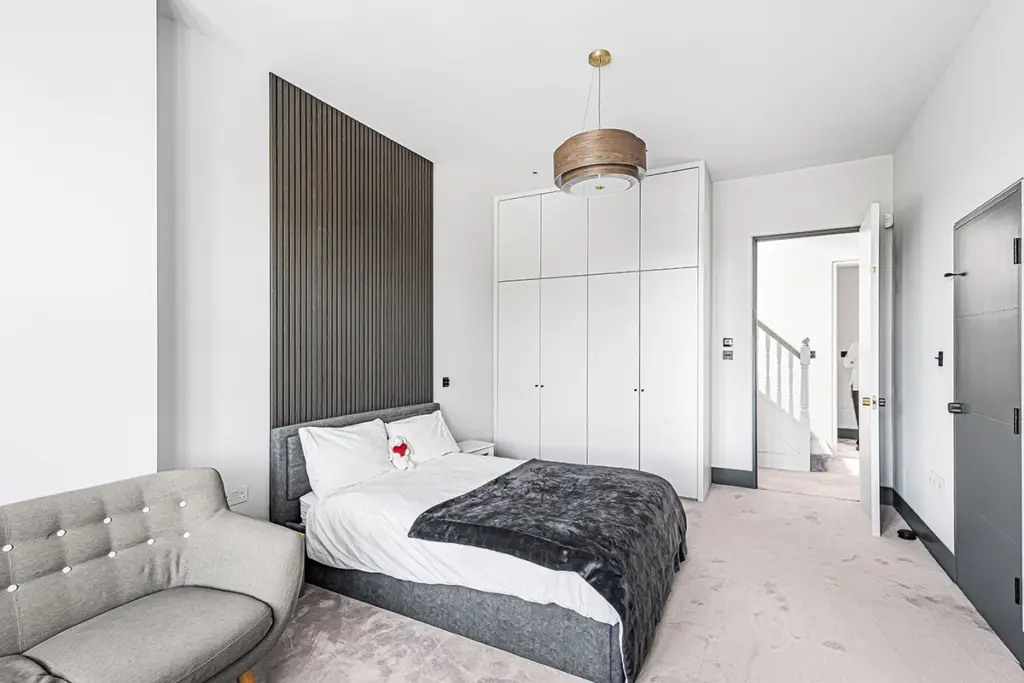 A bedroom featuring a bed with a dark grey headboard and matching base, fitted white wardrobes, a grey armchair, and a wooden pendant light.