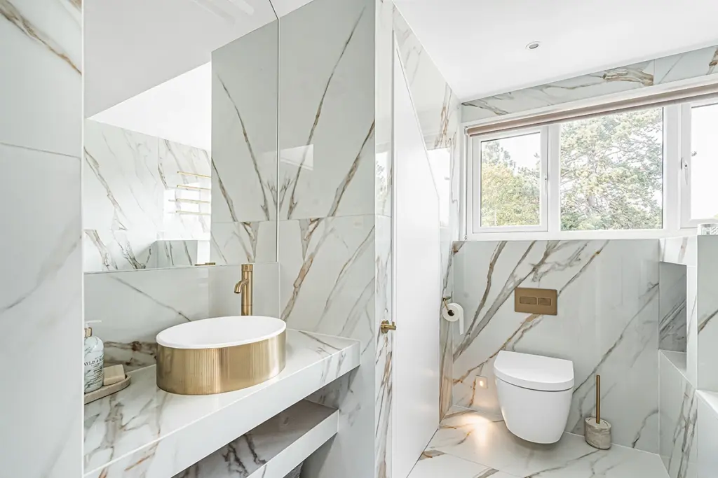 A modern bathroom with white marble walls, a gold vessel basin with a gold tap, and a wall-mounted toilet.
