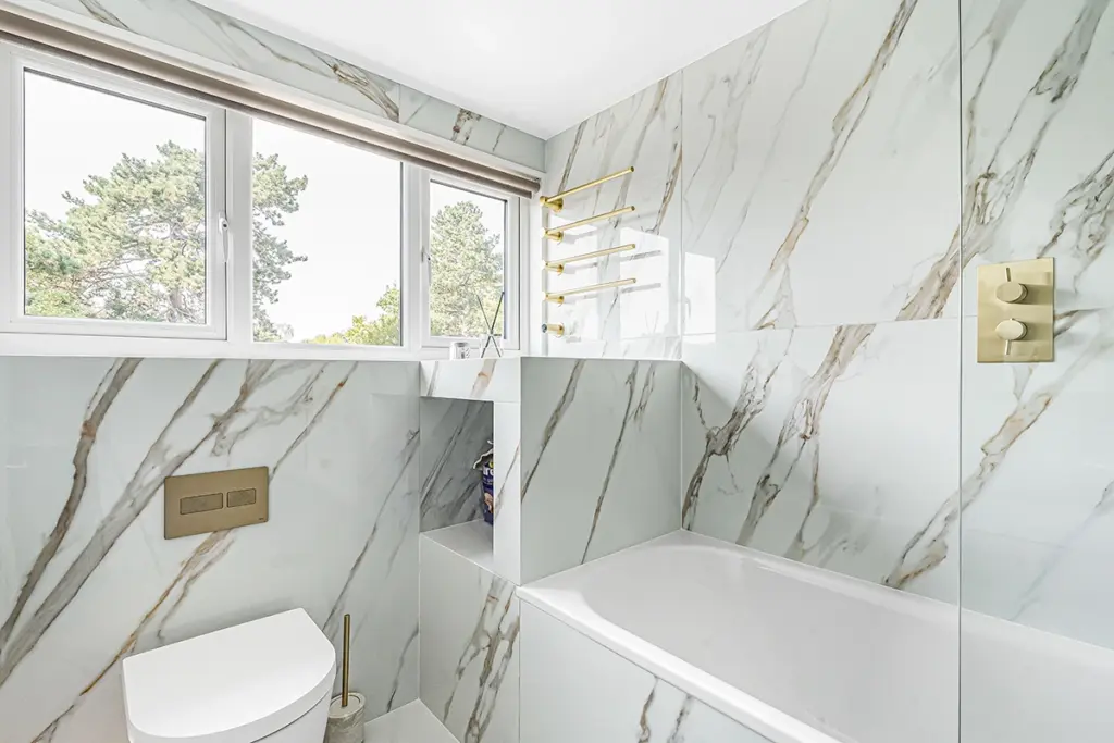 A bathroom featuring white marble-look wall tiles with gold veining, a white bath, a wall-hung toilet, and gold-finish fittings.