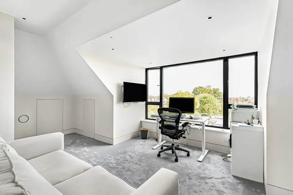 A modern home office in a bright loft room with a large desk, computer, ergonomic chair, and a white sofa, overlooking a view through a large window.