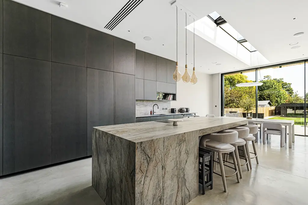 A modern kitchen featuring a large stone island with four bar stools, dark floor-to-ceiling cupboards, and a skylight above a dining area with a view of the back garden.