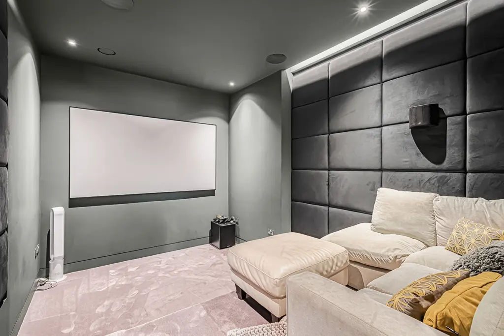 A home theatre room with a projection screen on a grey wall and a cream-coloured sectional sofa facing it against a dark, panelled wall.