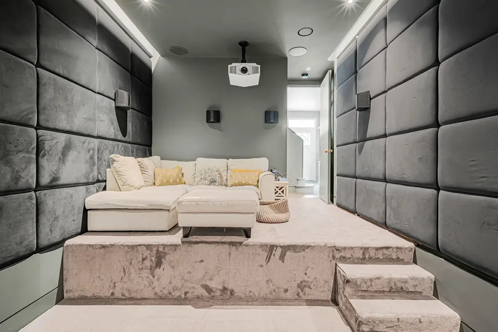 A home theatre room with a raised carpeted platform, a cream-coloured sectional sofa, and grey acoustic wall panels, featuring a ceiling-mounted projector.