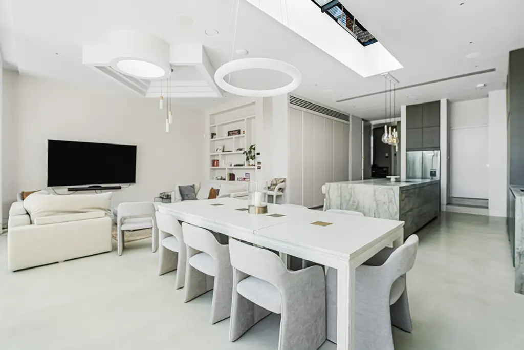 An open-plan living and dining room with a white dining table surrounded by six upholstered chairs, a white sofa facing a television, and a kitchen island with a stone worktop under a skylight.