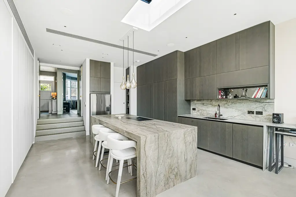 A modern kitchen featuring a stone-topped island with four white stools, dark grey cabinetry, and a view into a hallway with steps.