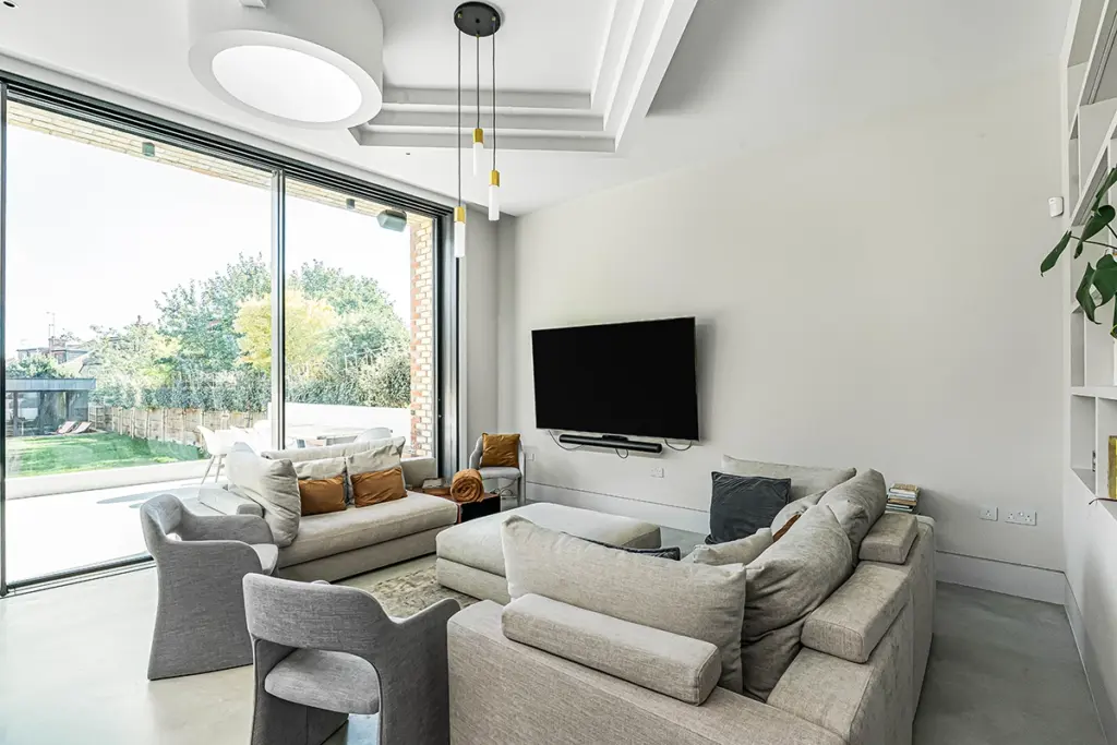 A modern living room with a large corner sofa, two armchairs, a wall-mounted television, and floor-to-ceiling glass doors opening to a garden.