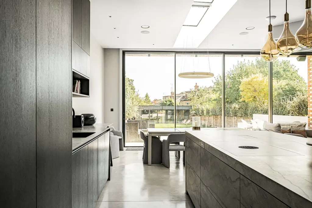 A modern kitchen with dark cabinetry, a large stone-topped island, and a dining area that opens to a garden through floor-to-ceiling glass doors.