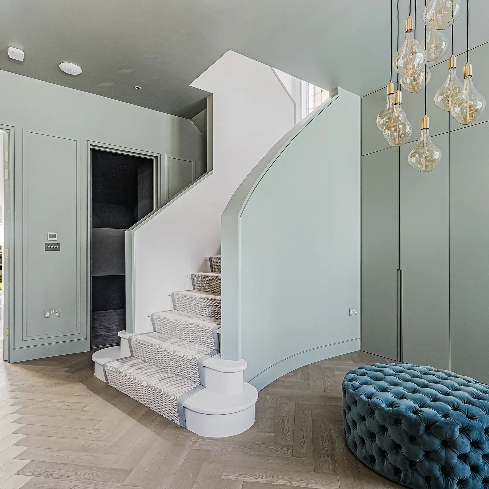 A curved staircase with a patterned runner, flanked by sage green walls and built-in cabinetry, with a blue tufted ottoman and a cluster of hanging light bulbs in the foreground.