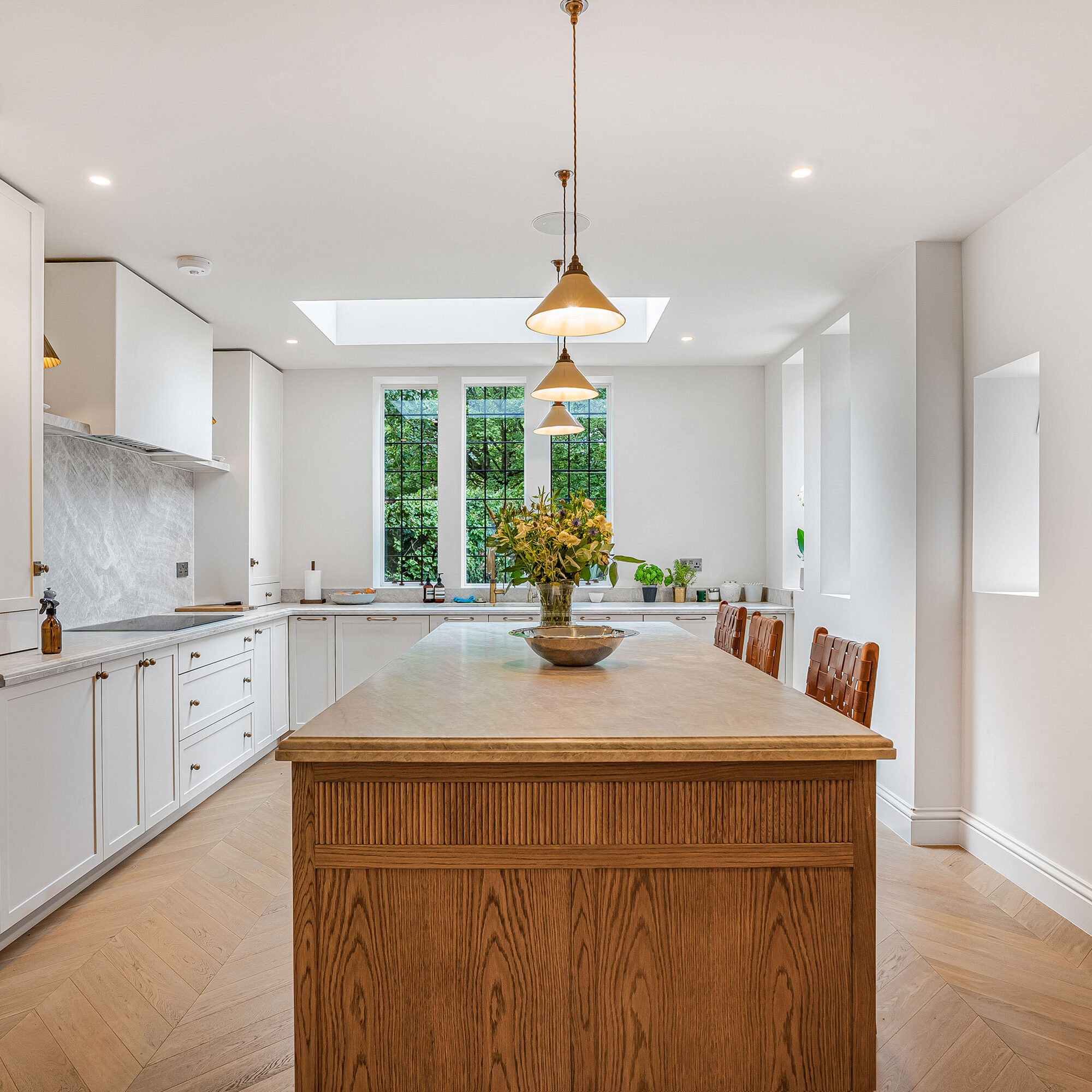 Bright modern kitchen with white cupboards, marble splashback, wooden island, pendant lights, and large windows. A vase with yellow flowers sits on the island, and wooden chairs line one side.