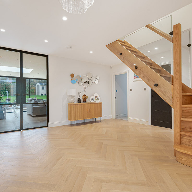 Spacious modern hallway with wooden herringbone floors, a wooden staircase with glass railing, a sideboard with decor, and glass doors leading to a bright living area and garden.